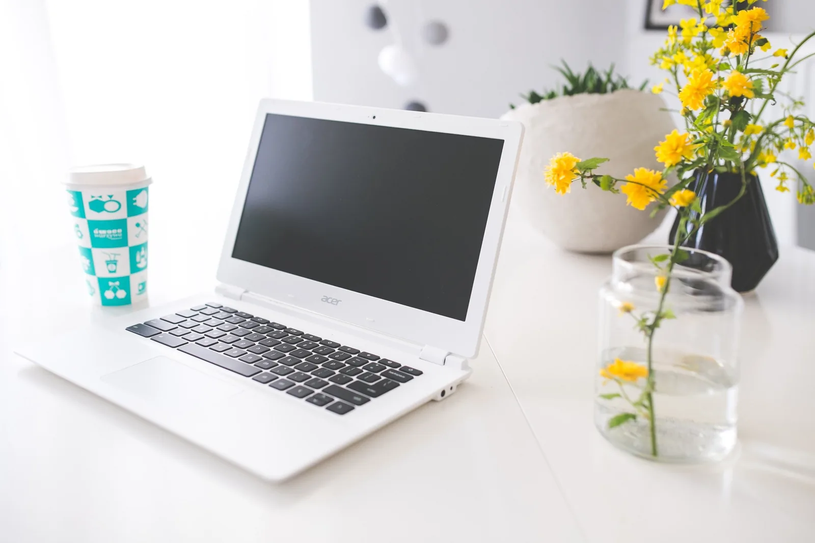Một laptop trắng đặt trên bàn cạnh cốc cà phê và hoa vàng trong góc làm việc sáng sủa.