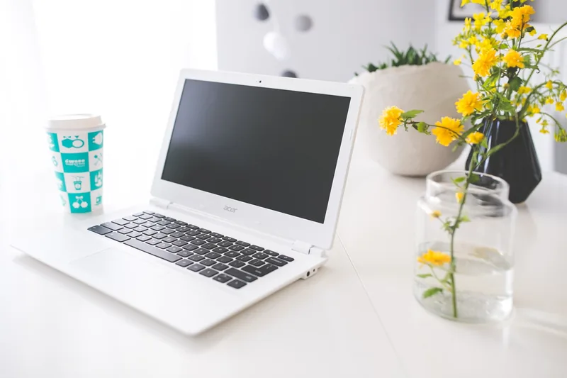 Một laptop trắng đặt trên bàn cạnh cốc cà phê và hoa vàng trong góc làm việc sáng sủa.