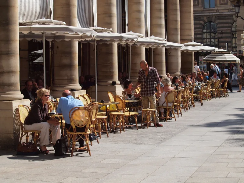 Khách dùng bữa tại hàng bàn cà phê ngoài trời dưới mái hiên sọc bên hàng cột ở Paris.
