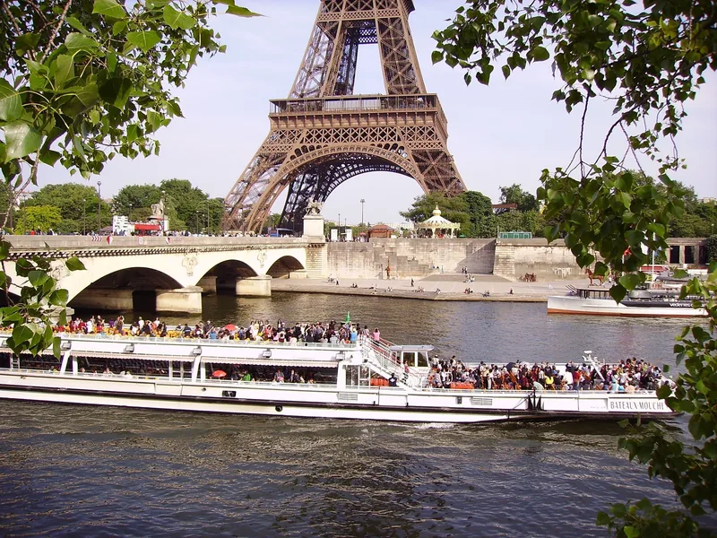 Một thuyền du lịch đông khách lướt trên sông Seine gần cầu, phía sau là tháp Eiffel sừng sững.