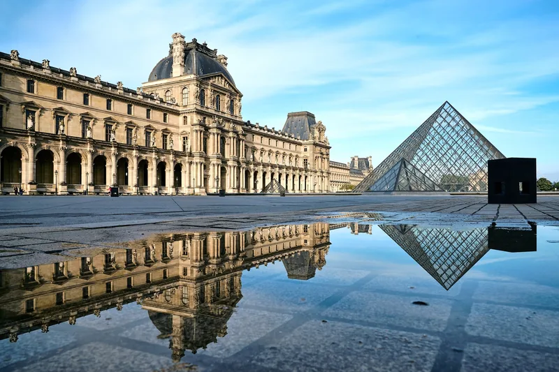 Kim tự tháp kính và mặt tiền cổ kính của bảo tàng Louvre phản chiếu trên vũng nước dưới bầu trời trong xanh ở Paris.