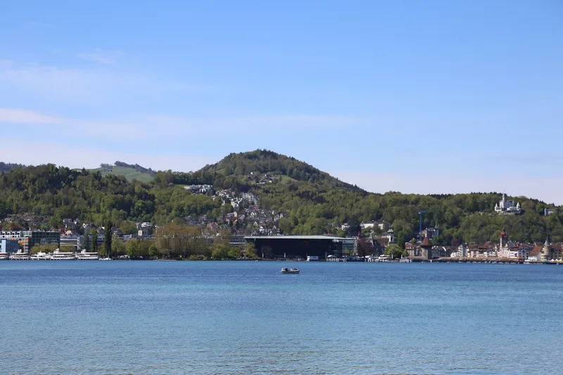 Mặt hồ phẳng lặng trải dài hướng về những ngọn đồi xanh của Lucerne, cùng các tòa nhà ven hồ và một chiếc thuyền nhỏ.
