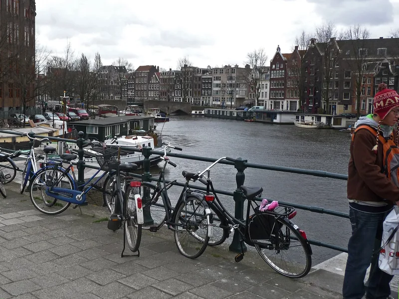 Những chiếc xe đạp đậu dọc hàng rào bên kênh đào Amsterdam, với thuyền nhà và nhà phố mái nhọn phía xa.