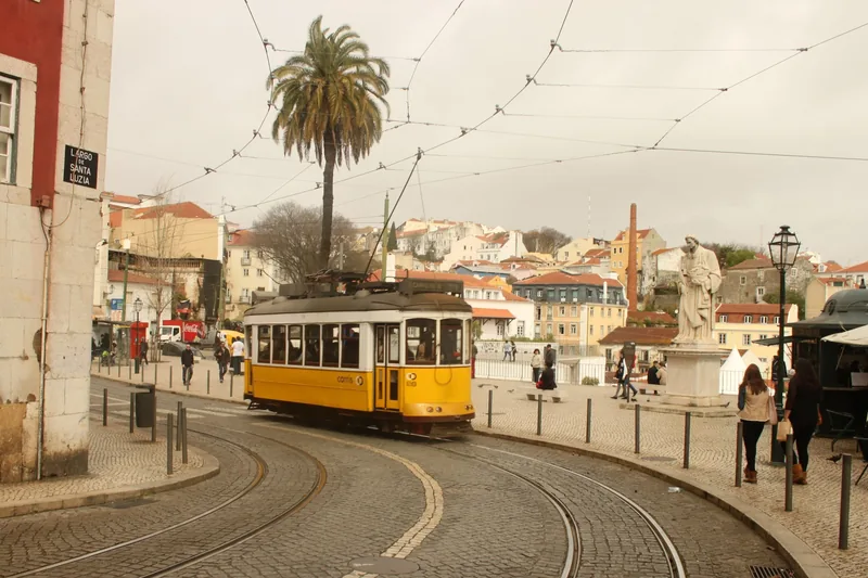 Xe điện vàng cổ điển chạy trên đường ray lát đá tại Largo de Santa Luzia ở Lisbon, gần bức tượng và cây cọ.