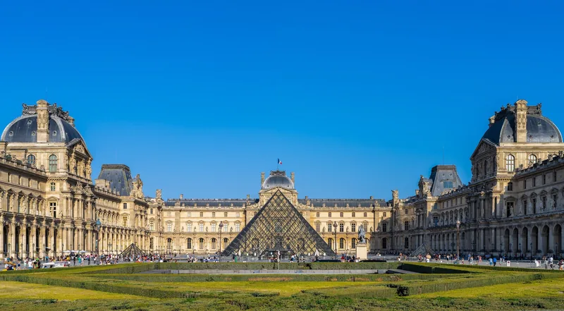 Kim tự tháp kính của Bảo tàng Louvre nổi bật giữa các cánh cung điện cổ kính dưới bầu trời xanh ở Paris.