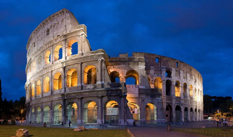 Đấu trường Colosseum cổ đại ở Rome sáng rực trong ánh đèn vàng ấm dưới bầu trời xanh thẫm lúc chạng vạng.