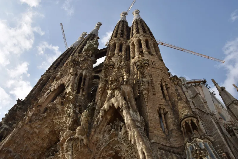 Các tháp nhọn và họa tiết đá chạm khắc tinh xảo của Sagrada Familia vươn lên trời xanh ở Barcelona, có cần cẩu xây dựng.